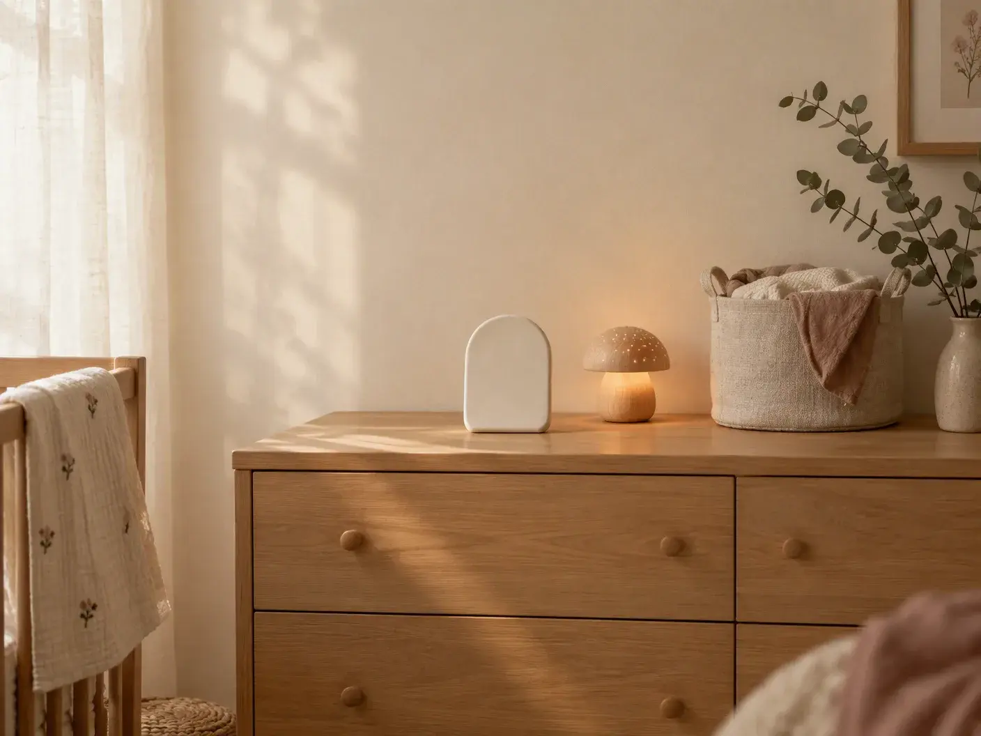 A light oak dresser against a cream wall with a small cream plaque and a warmly glowing wooden night light, a linen basket and a sprig of eucalyptus
