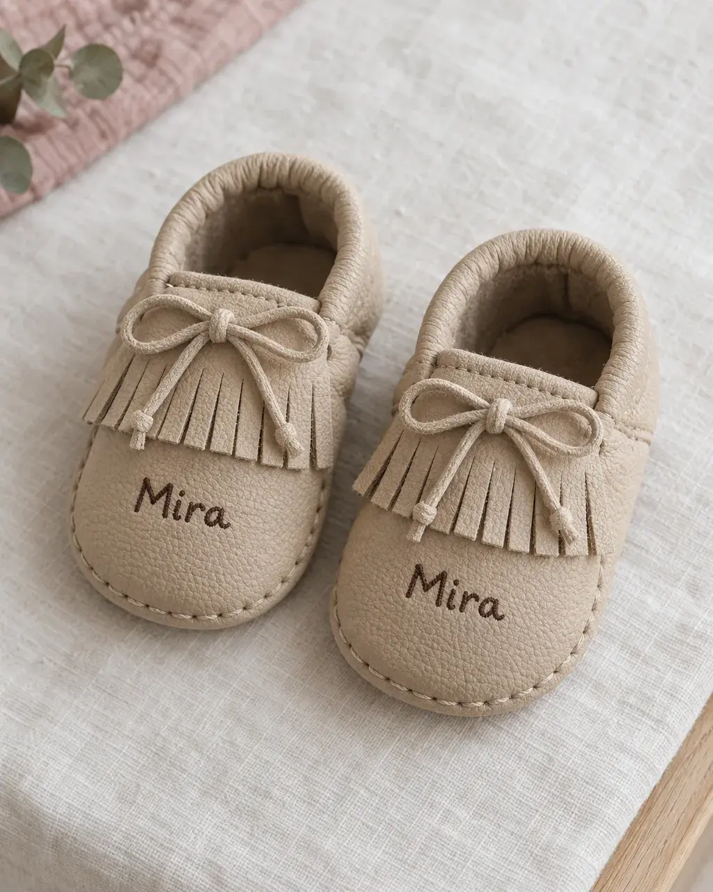 A pair of sand-coloured fringed baby shoes with Mira written on both toes